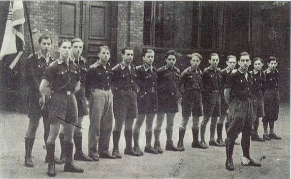 młodzi mężczyźni w strojach przypominających mundury stoją w szeregu po sztandarem
